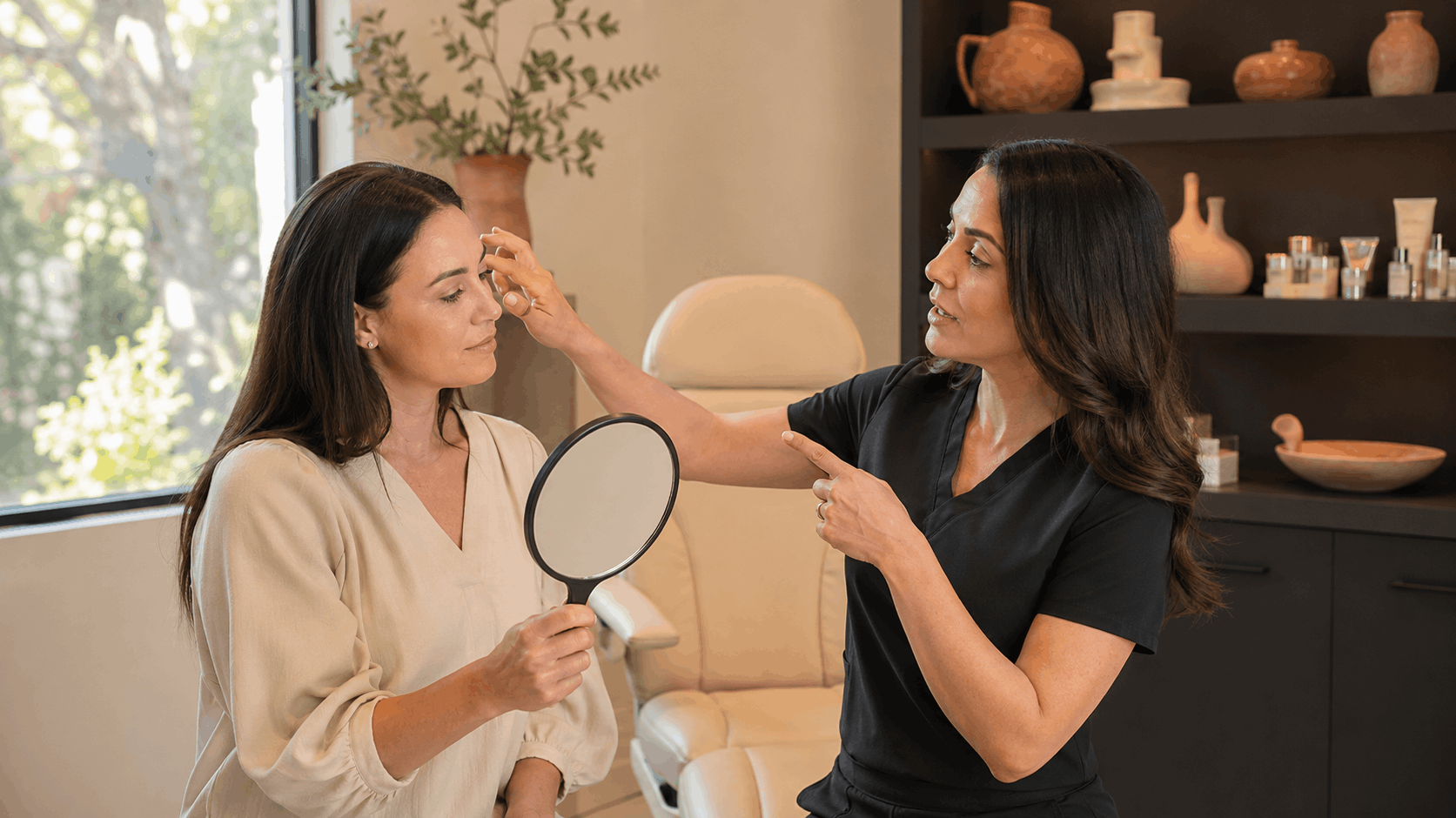 provider examining a patient's forehead while discussing cosmetic treatment options with a handheld mirror.