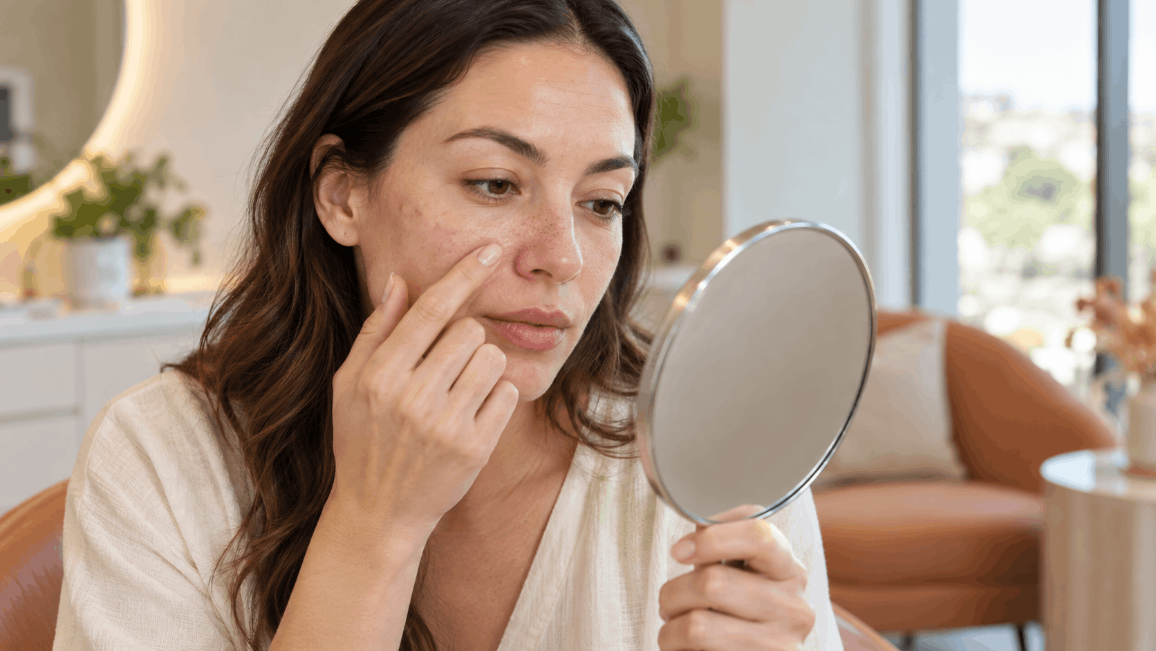 woman examining visible pores and skin texture on her cheek in a handheld mirror.