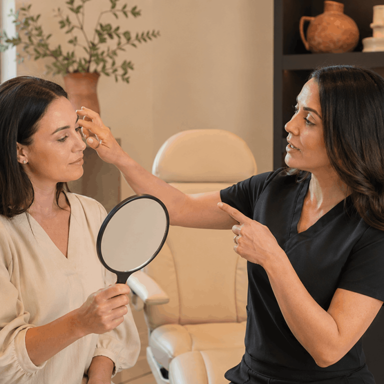 Provider examining a patient's forehead while discussing cosmetic treatment options with a handheld mirror.